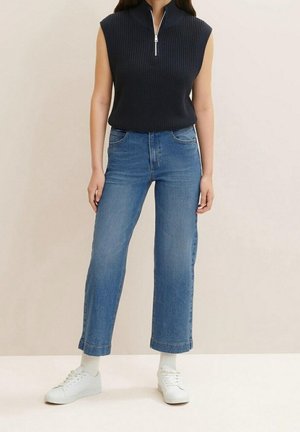 Persoon met mouwloos zwart gebreid topje met ritssluiting aan de kraag, blauwe jeans met rechte pijpen, witte sokken en witte sneakers, staand tegen een beige achtergrond.