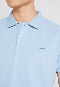 Polo bleu clair en tissu texturé, avec une patte de boutonnage à deux boutons et un petit logo "ESPRIT" brodé sur la poitrine.