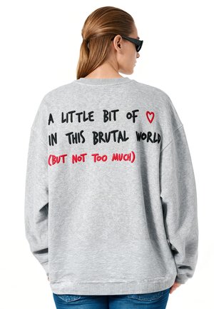 Personne aux longs cheveux attachés, portant des lunettes de soleil et un sweat-shirt gris avec un texte noir et rouge dans le dos.