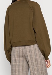 Olivově zelená volná mikina s nízko posazenými rameny a žebrovaným lemem, kombinovaná s károvanými kalhotami v béžové a černé barvě.