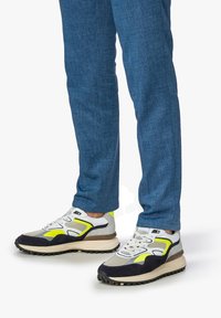 Scarpe sportive multicolori con una tomaia in mesh e accentuate da dettagli grigi, bianchi e neri, con una suola testurizzata, indossate con pantaloni blu.