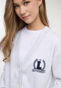 Cardigan grigio chiaro con collo a coste e chiusura con bottoni. Presenta un logo ricamato in blu navy con la lettera "L" e la scritta "ST LAS VEGAS" sul petto.