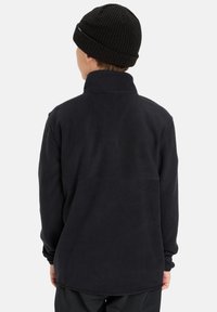 Schwarzer Fleece-Pullover mit hohem Kragen und langen Ärmeln. Weiche Textur und schlichtes Design. Getragen mit einer schwarzen Strickmütze.