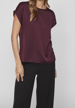 Femme portant un chemisier violet foncé à manches courtes avec des revers et un pantalon noir, debout devant un fond gris clair uni.