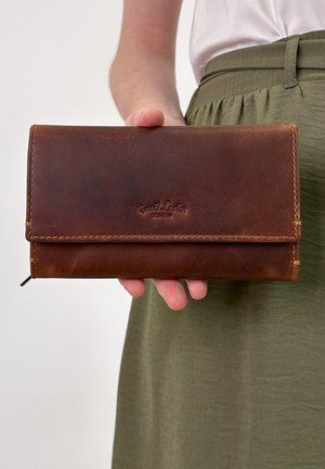 Main tenant une portefeuille en cuir marron avec des détails de couture, sur un fond de haut blanc et de jupe olive.