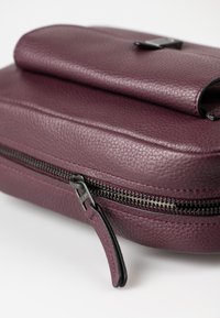 Tief lila Lederhandtasche mit einer Reißverschlussöffnung und einer flachen Klappe oben. Sie verfügt über eine strukturierten Oberfläche und schwarze Hardware-Akzente.