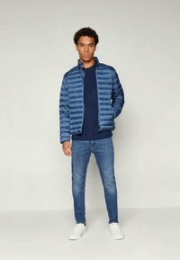 Chaqueta de plumas azul con rayas horizontales, con un cuello alzado, combinada con un suéter azul marino y vaqueros azules. El modelo lleva zapatillas blancas.