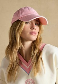 Joven mujer con cabello largo y rubio, lleva una gorra de béisbol rosa y un suéter blanco con rayas en V rosas y azules.