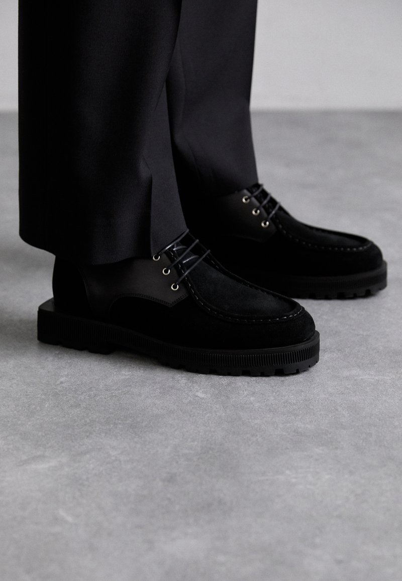 Melni apavi, kas izgatavoti no zamša un gludas ādas kombinācijas, ar biezu gumijas zoli un izteiktiem šuvju dekoriem.