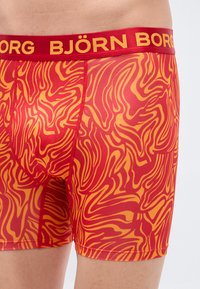 Κόκκινο και κίτρινο ανδρικό εσώρουχο μποξέρ με ευρεία κόκκινη ζώνη μέσης που φέρει την ένδειξη "BJÖRN BORG" με κίτρινα γράμματα, κατασκευασμένο από λείο, ελαστικό ύφασμα.