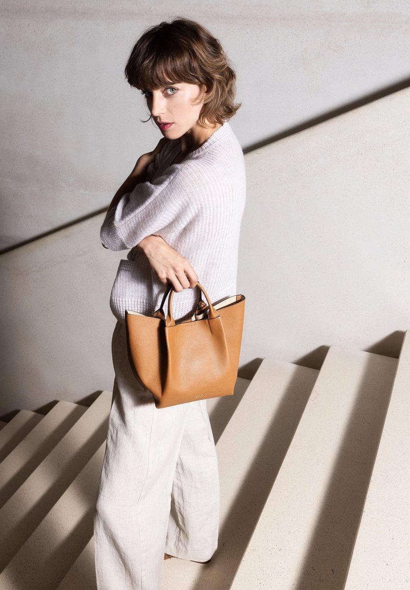 Handtasche aus cognacfarbenem Leder mit abgerundeten Seiten, zwei kurzen Griffen und minimalistischem Design. Modell trägt einen leichten Pullover und weit geschnittene Hosen auf einer Treppe.