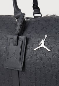 Schwarze Duffel-Tasche aus strukturiertem synthetischem Material, mit einem erhabenen Muster, silbernen Beschlägen und einem weißen Jumpman-Logo-Akzent.