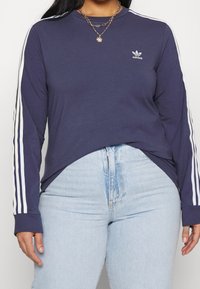 Námořnická košile s dlouhým rukávem a bílým logem Adidas, s bílými vertikálními pruhy na rukávech. V kombinaci se světle modrými džíny s vysokým pasem.