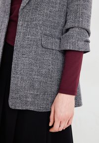 Sivi teksturirani sako s podvrnutim rukavima, sa malim džepom, nosi se preko maroon dugih rukava. Crna tkanina vidljiva ispod.