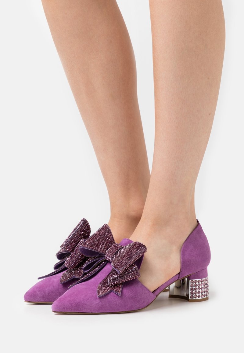 Chaussures basses en daim violet avec de grands nœuds texturés et des ornements en strass sur des talons carrés, portées pieds nus.