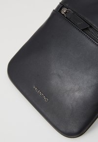 Juodas odinis krepšys su lygaus paviršiaus, minimalistiniu dizainu, su užtrauktuku ir sidabriniu "VALENTINO" logotipu priekyje.
