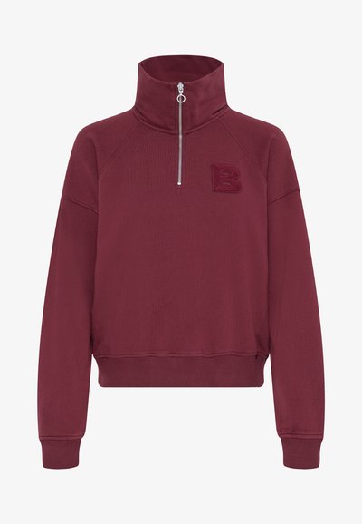 Burgundska zip-upbluza s visokim ovratnikom, izvezenim slovom "B" i rebrastim manžetama i donjim rubom, napravljena od mekanog materijala sa glatkom teksturom.