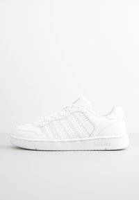 Bílé sportovní boty s perforovanými detaily, plochými tkaničkami, zaoblenou špičkou a polstrovanou podrážkou. Na boku je logo K-Swiss.