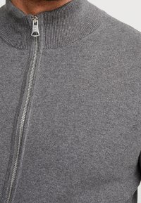 Grå zip-up-tröja med hög krage, med en metalldragkedja och ribbad textur. Slät tyg med ett subtilt, jämnt mönster.