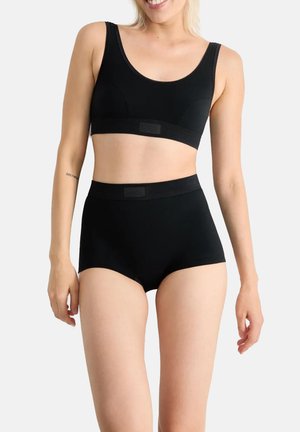 Frau trägt schwarzen nahtlosen Sport-BH und hoch taillierte Unterwäsche, lächelt, helle Haut, steht vor neutralem weißen Hintergrund.