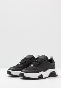 Pair of black and white Versace Jeans Couture chunky sneakers with laces and branded rubber soles on a white surface.