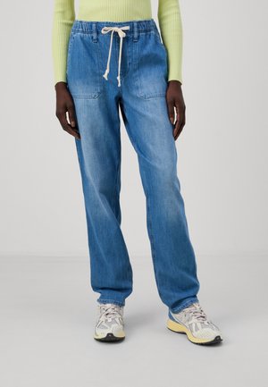 Personne portant un pantalon en denim bleu clair à cordon de serrage, des baskets blanches et grises, et un haut à manches longues côtelé vert clair, debout sur un fond neutre.