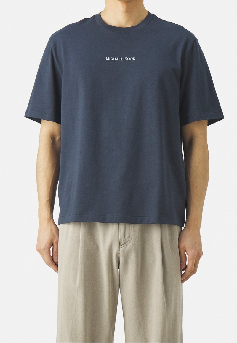 T-shirt in cotone blu navy con maniche corte, scollatura rotonda e testo bianco "MICHAEL KORS" centrato sul petto. Texture liscia.