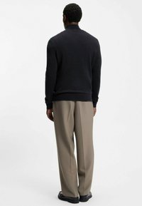 Pull noir à col montant, associé à un pantalon beige. Le tissu semble lisse et la coupe est décontractée. Silhouette angulaire et minimaliste.