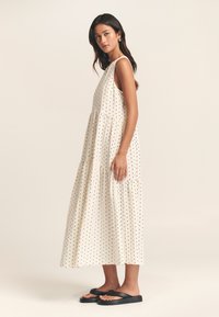 Robe sans manches blanche avec des pois noirs, design en couches, tissu fluide, portée avec des tongs noires, capturée de côté.