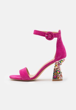 Pink ruskindsandal med høje hæle, ankelrem og en dekoreret chunky hæl med flerfarvede ædelstensdetaljer.