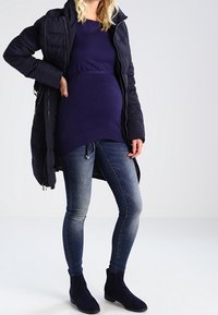 Pitkä tummansininen toppatakki tumma violetti paita päällä, yhdistettynä sinisiin kapeisiin farkkuihin ja tummansinisiin nilkkasaappaisiin. Mukana vetoketju ja kiristysnauha.