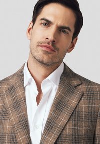 Brązowa kratkowana marynarka z niebieskimi akcentami, teksturowany materiał, klapy w kształcie notcha oraz biała koszula o kołnierzyku pod spodem. Gładkie tło.