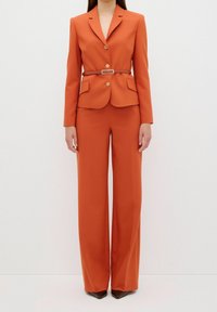 Femme portant un costume orange sur mesure avec un blazer croisé, un pantalon large assorti et une ceinture marron à la taille.