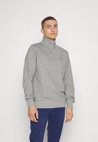 Felpa grigia con zip e colletto alto, polsini a coste e un piccolo logo ricamato sul petto, realizzata in materiale morbido. Abbinata a pantaloni blu.