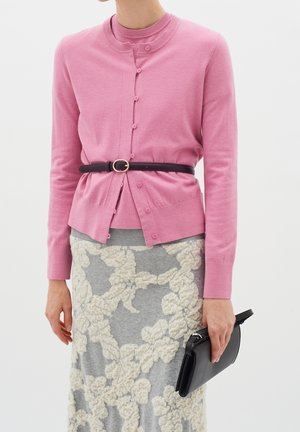 Femme portant un cardigan rose boutonné cintré avec une ceinture noire, une jupe grise avec un motif floral blanc texturé, tenant une pochette noire.