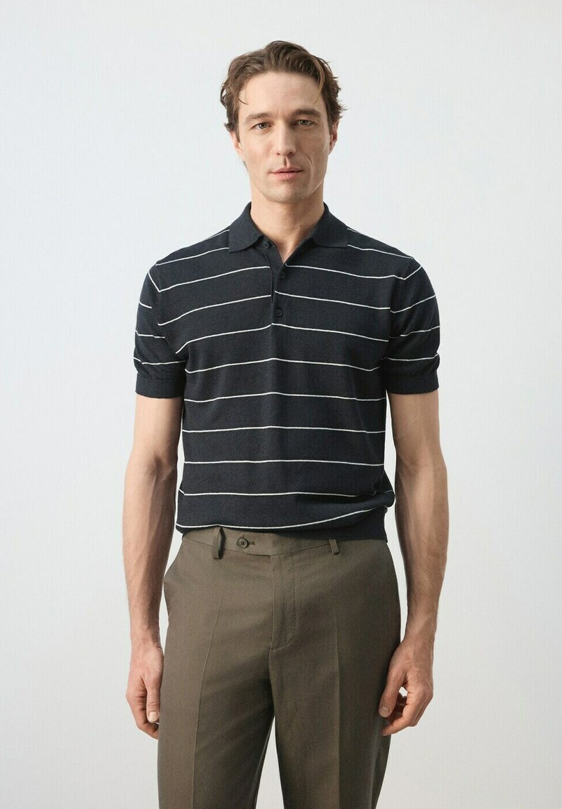 Homme portant un polo à manches courtes noir avec des rayures horizontales blanches et un pantalon vert olive, debout devant un fond clair uni.