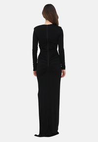 Femme aux longs cheveux bruns portant une robe noire cintrée, à manches longues, longueur au sol, debout, dos tourné vers l’avant.