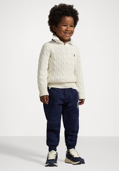 Vit kabelstickad tröja, marinblå sweatpants med en broderad design, och krämfärgade/mörka sneakers. Kragen är lager-på-lager med en vit skjorta under.