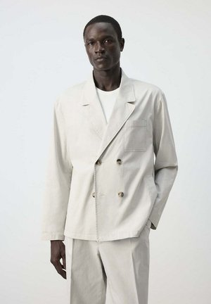 Hombre alto que lleva un blazer cruzado beige claro y pantalones a juego sobre un fondo liso.