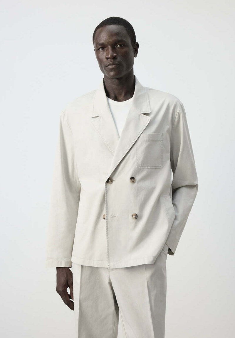 Homme grand portant un blazer croisé beige clair et un pantalon assorti sur un fond uni.