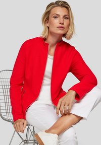 Red zip-up jacket with a textured design, featuring a high collar and two side pockets, paired with a white top and pants.