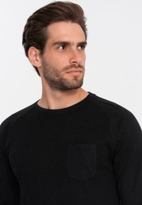 Musta pitkähihainen paita, jossa on ribbitekstuuri, pyöreä kaula-aukko ja pieni rintatasku. Sujuva kangas, jossa on kontrastiväriset hihadetailit.