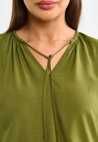 Blouse verte avec un col en V et des détails froncés. Le tissu a une texture lisse, avec un accent noué au niveau du col.