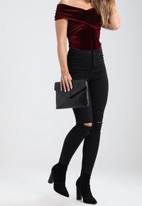 Sametová off-shoulder blůza v burgundské barvě, černé skinny džíny s vysokým pasem a vytrženými koleny, černá kabelka do ruky a černé kotníkové boty na podpatku.