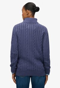 Lila Strickpullover mit Zopfmuster und hohem Kragen. Verfügt über gerippte Kanten und eine lockere Passform. Kombiniert mit blauen Jeans für einen Kontrast.