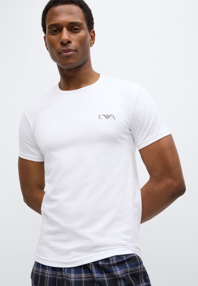 Bílé tričko s krátkým rukávem a kulatým výstřihem, s malým černým logem na levé části hrudi. Tkanina se zdá být hladká a lehká.