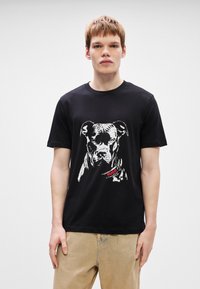 T-shirt de algodão preto com um gráfico de um cão branco e o texto "HUGO". Mangas curtas e gola redonda, ajuste descontraído. Fundo liso.