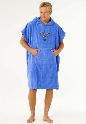 Ung mand iført en blå hættehåndklædeponcho med stor frontlomme og "Rip Curl"-logo, stående barfodet på en hvid baggrund.