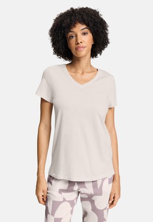 Donna con capelli ricci che indossa una maglietta semplice beige chiaro a maniche corte con scollo a V e pantaloni fantasia con forme astratte.