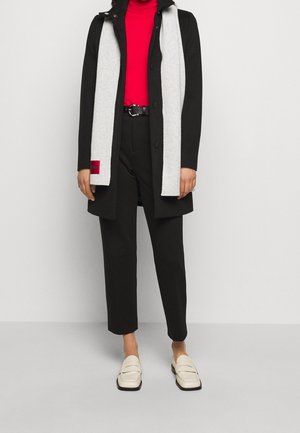 Personne portant un manteau noir, une écharpe grise, un col roulé rouge, un pantalon noir, une ceinture noire et des mocassins blancs, debout devant un fond clair uni.